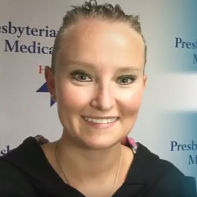 Jessica Barton smiles while wearing a black hoodie and is standing in front of a banner with the Presbyterian/St. Luke's Medical Center logo.