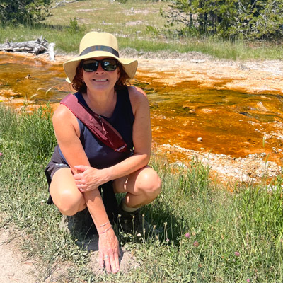 Leslee kneels down to touch the ground outdoors, while wearing a sunhat, sunglasses, and a bag over her shoulder.