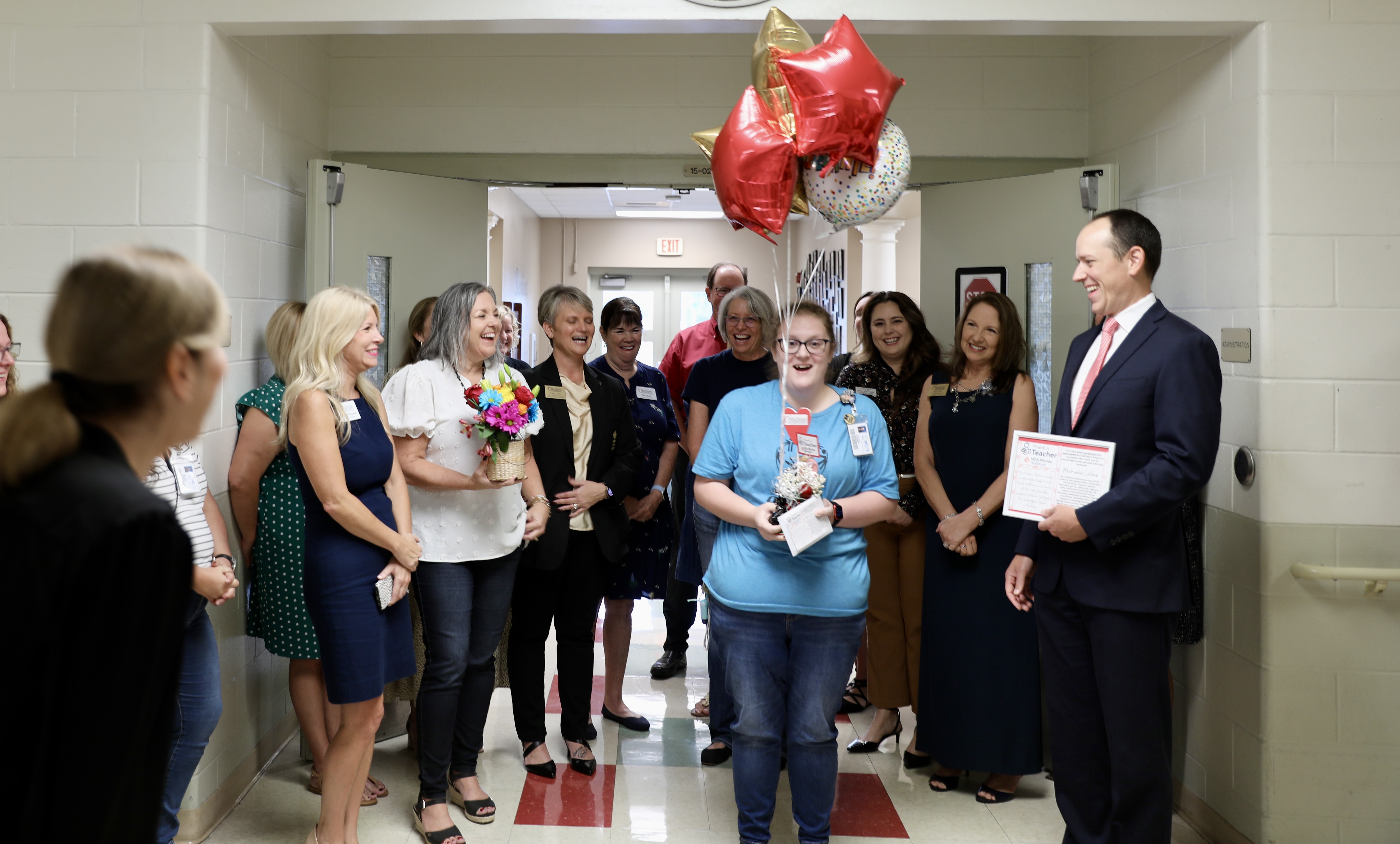 A group of people standing in a hallway in a school. One woman is holding balloons, one woman is holding flowers and a man in a suit is holding a plaque.