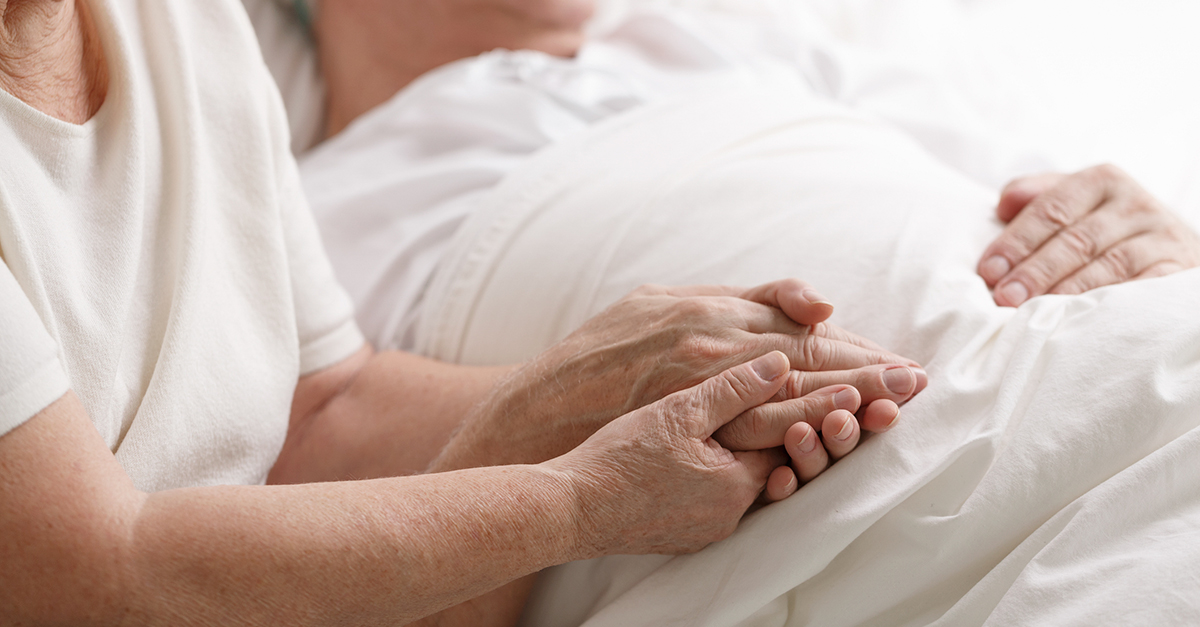 A senior woman holds her husband's hand, while he's lying in a patient bed.