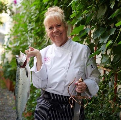Susie Von Willer Linford posing in her chef uniform, while holding a fish and a sword.