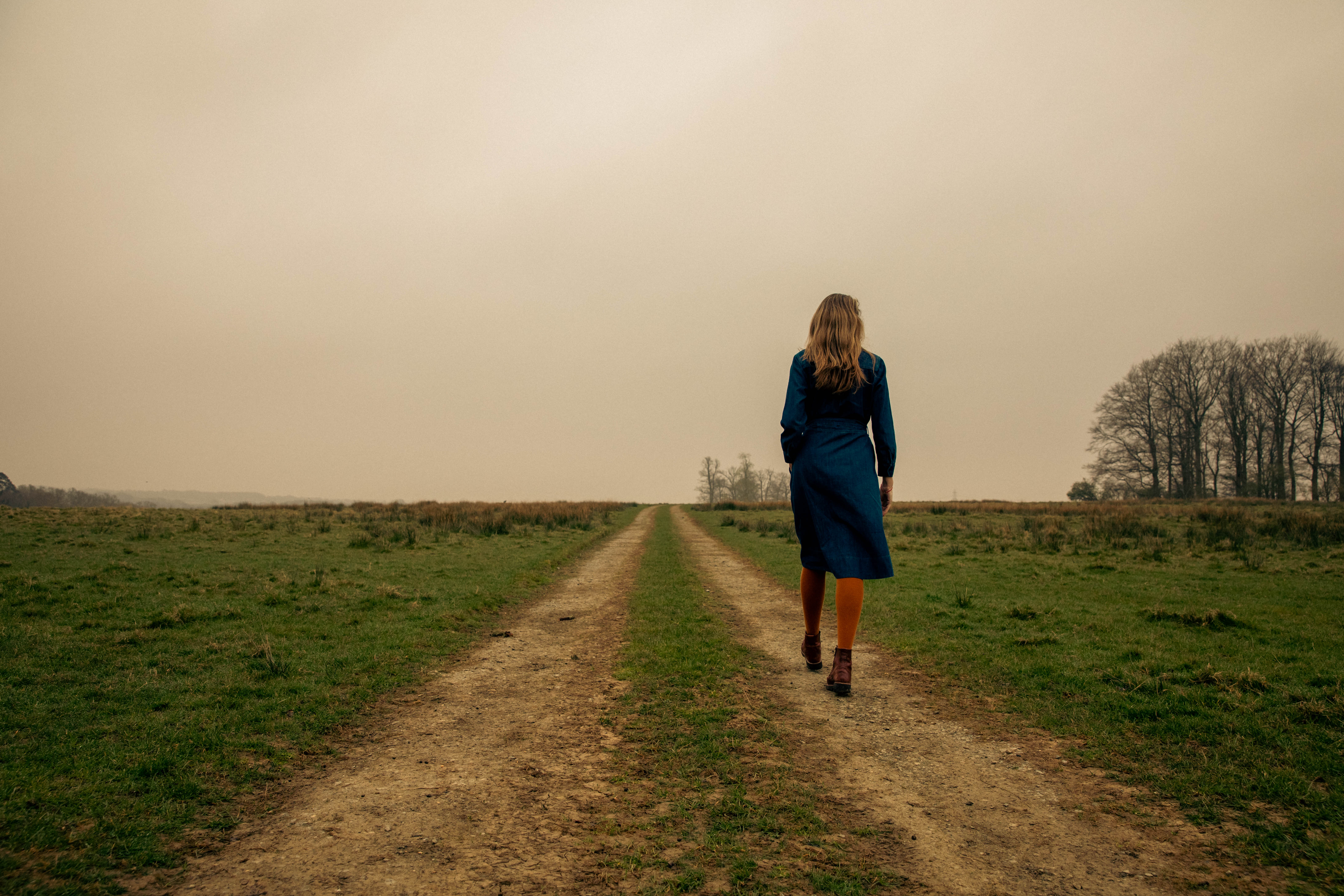 Woman in blue dress  walking down dirt path in grass field on a gray fall day