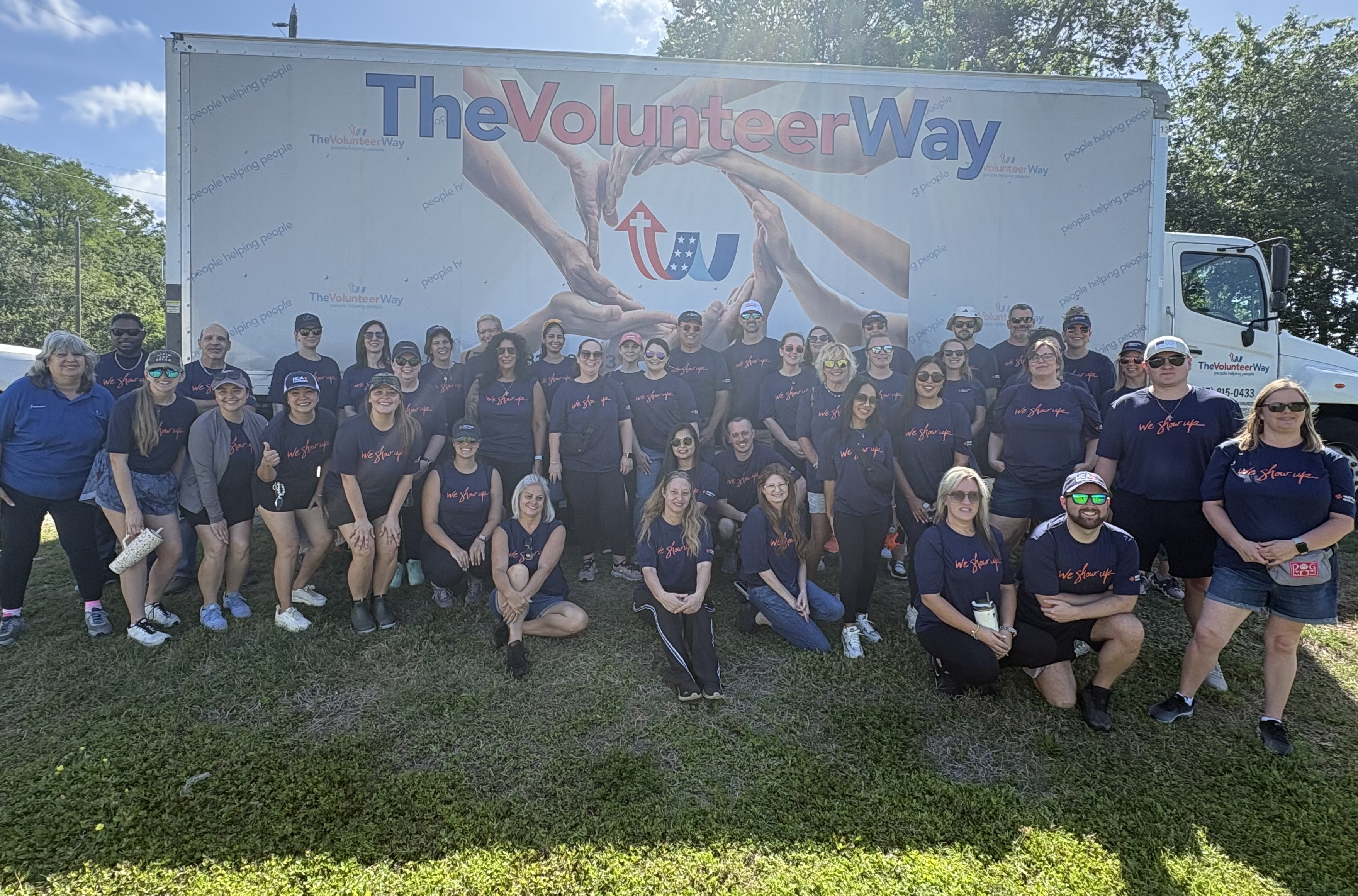 Group of hospital and community volunteers standing outdoors in front of a truck displaying a large ‘The Volunteer Way’ banner during a volunteer event.