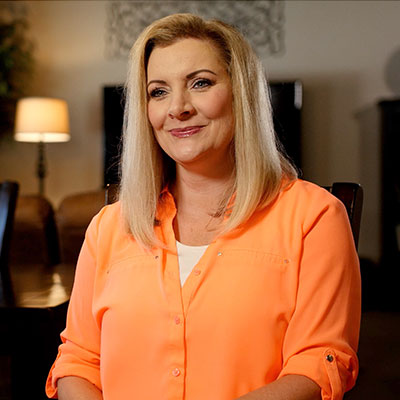 Brenda is seated in an orange button down t-shirt in an interior setting. 
