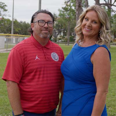 Two people standing together outdoors on a grassy area near a sidewalk, with trees, a yellow fence, and buildings in the background. One person is wearing a red sports polo shirt, and the other is wearing a bright blue dress.