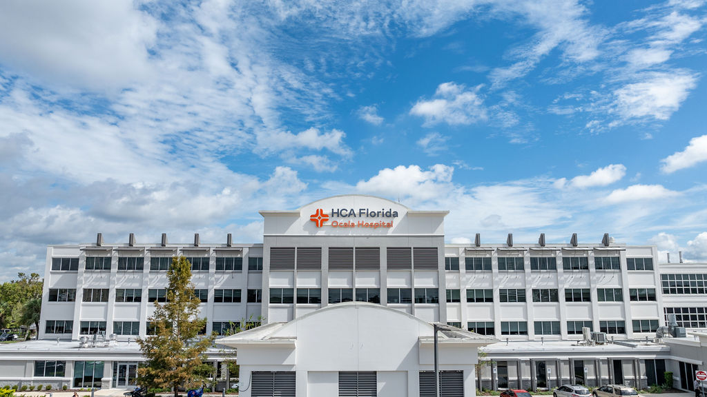 Front view of HCA Florida Ocala Hospital, a multi-story white medical building with a central entrance and signage under a blue sky with scattered clouds.