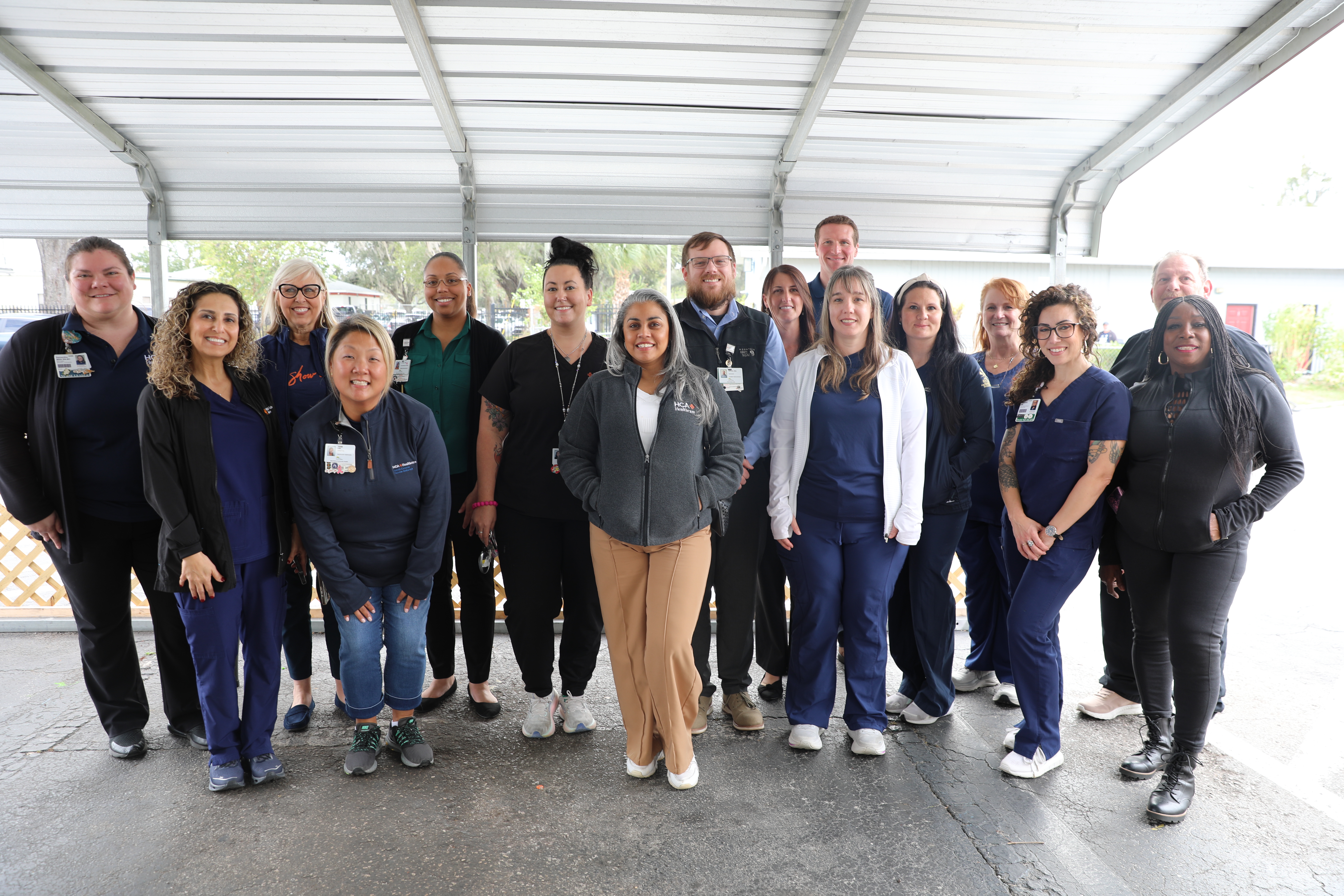 A group of 13 women and three men, dressed in either hospital scrubs or business clothes, stand in a row and smile while posing.