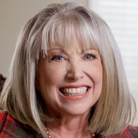 Dee smiles while wearing a red and green plaid cardigan.
