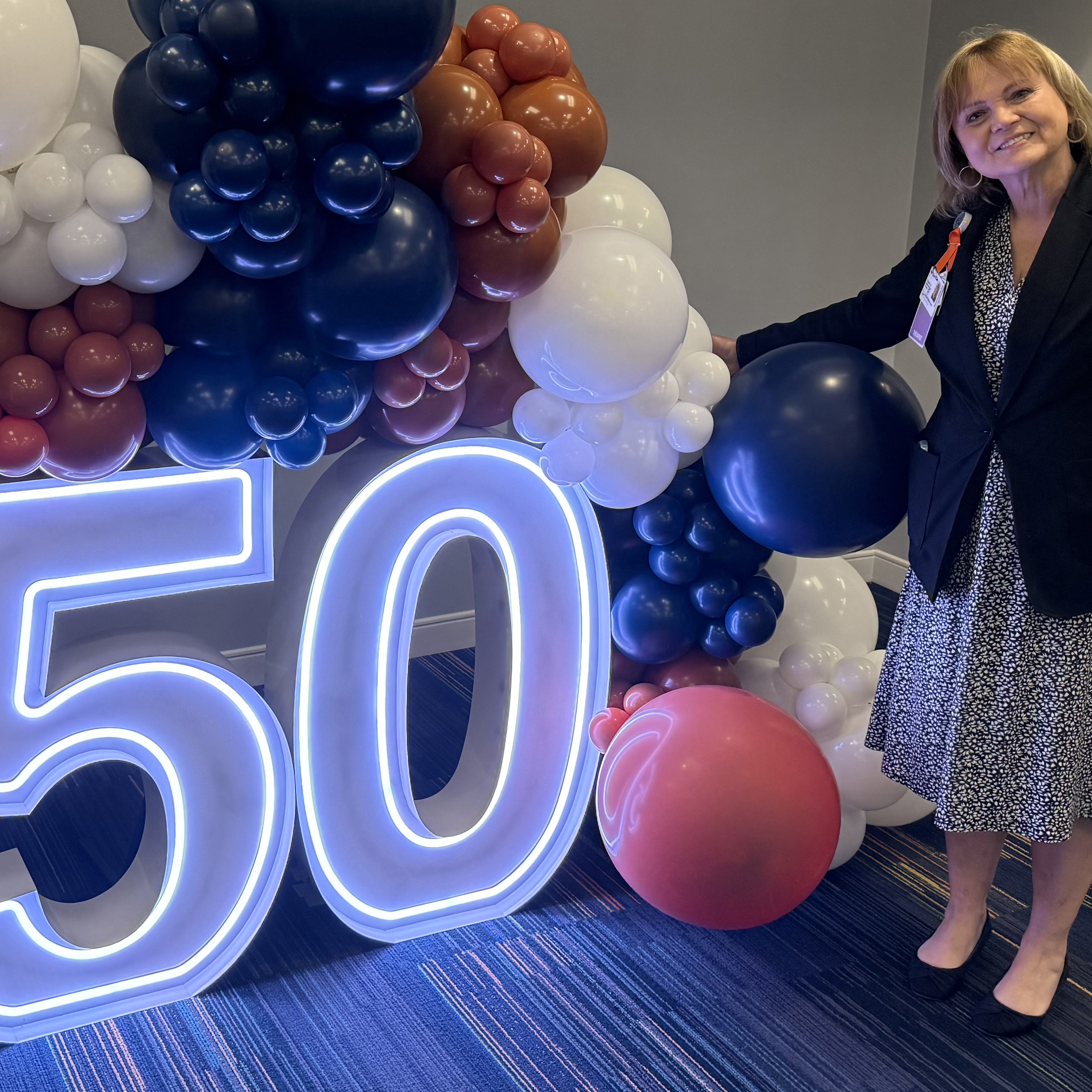 Lighted sign that with the letters 50. The sign is surrounded by balloons. A women with brown hair, wearing a skirt, stands to the right of the sign. 