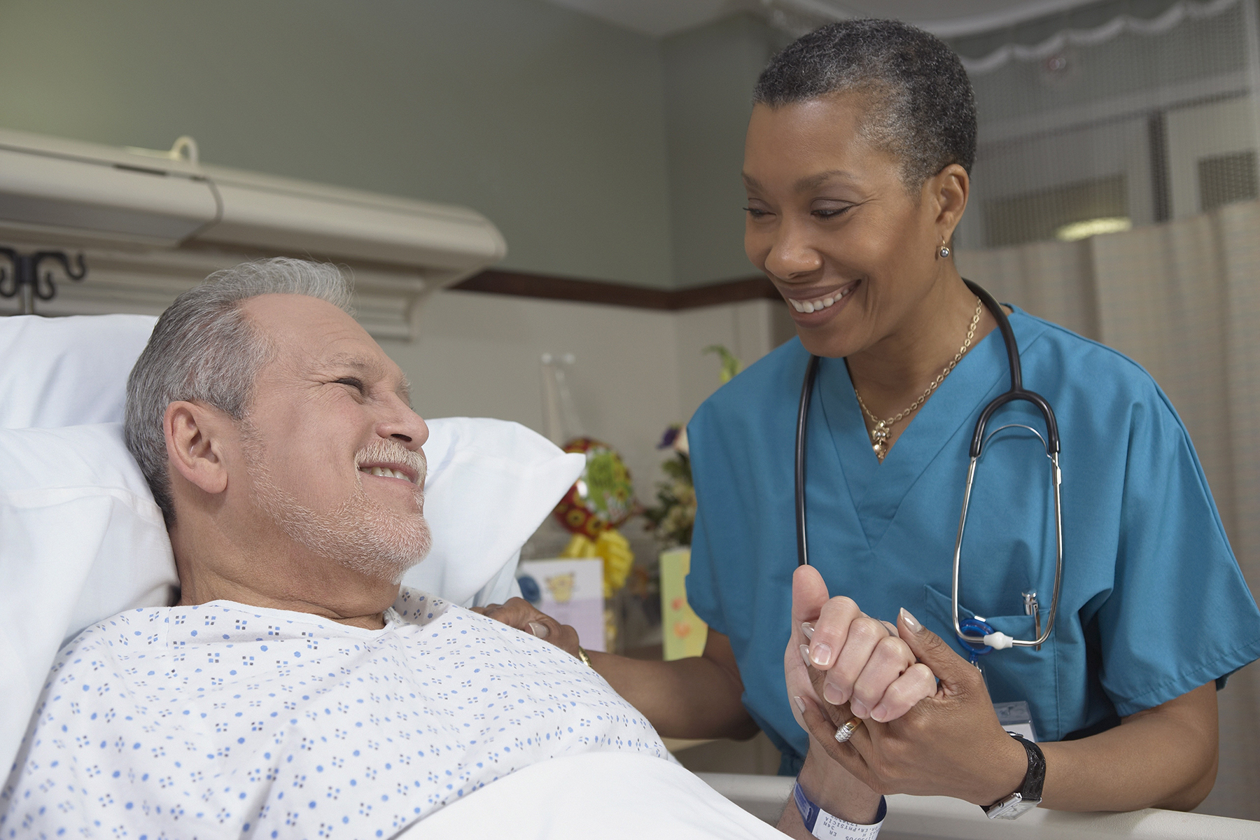 A female nurse smiling and holding the hand of her senior male patient, who is lying in bed.