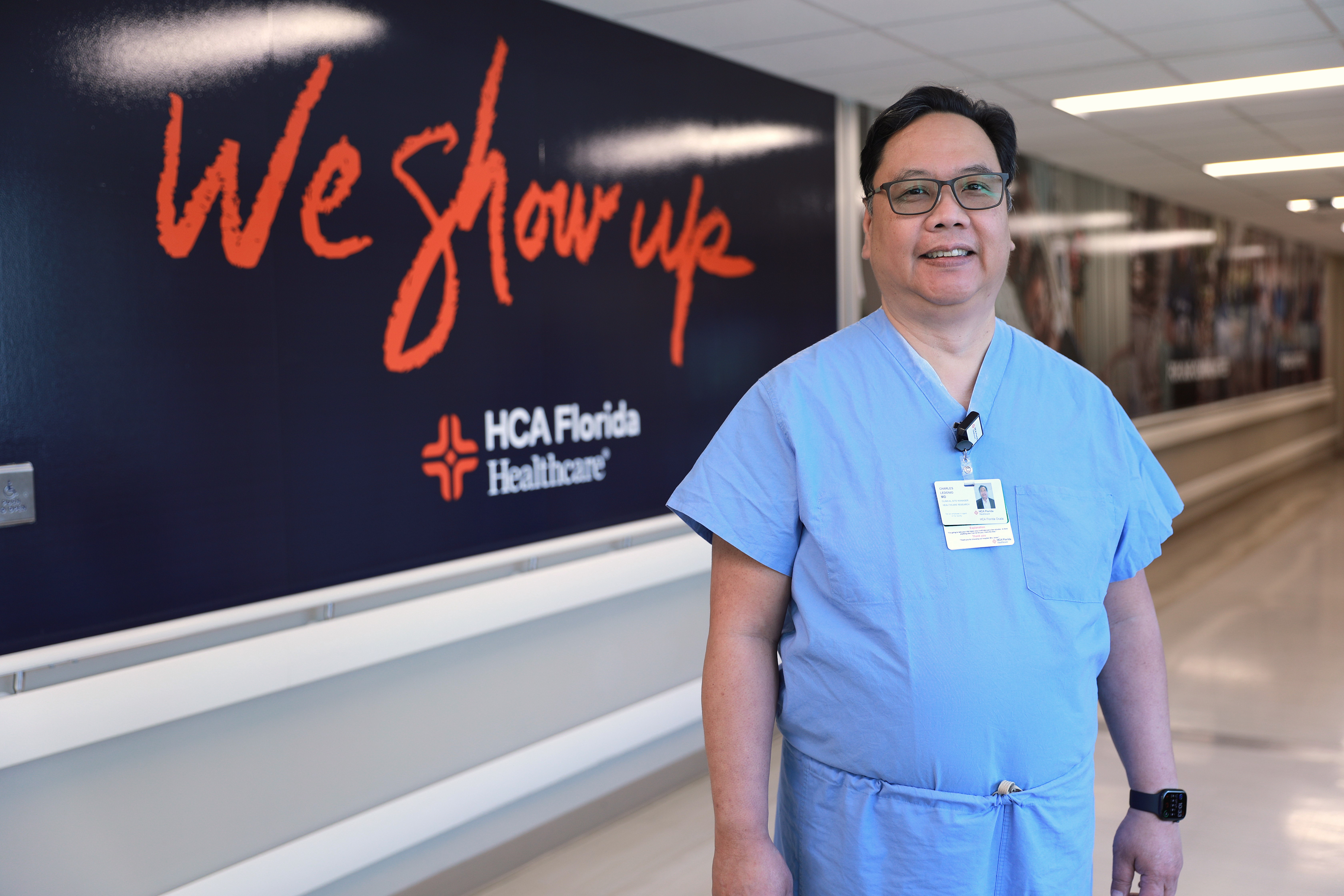 Dr. Charles Ledonio wearing hospital scrubs standing in a hospital hallway in front a We Show Up® mural.