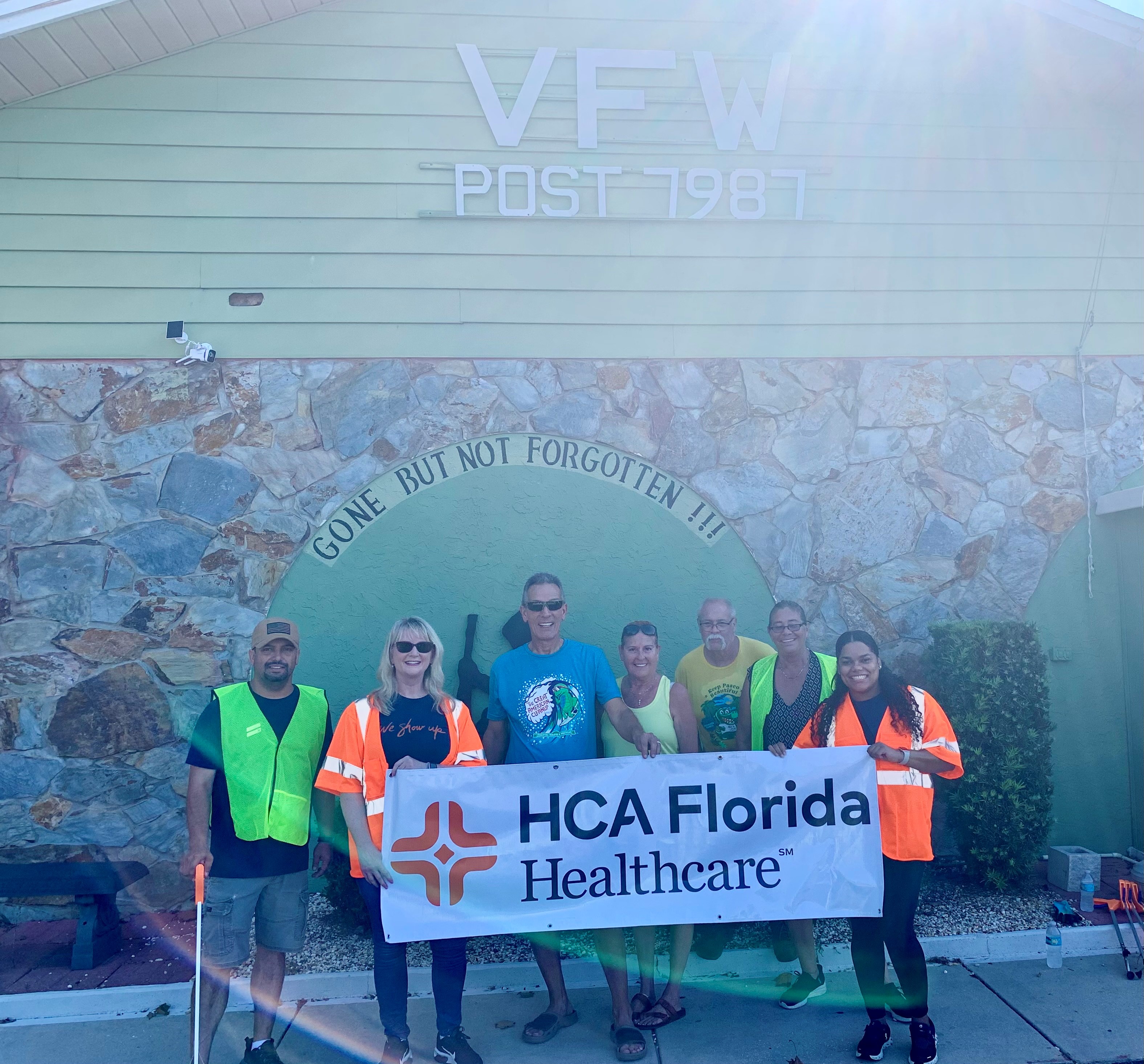 HCA Florida Trinity Hospital volunteers holding HCA Healthcare banner, standing in front of VFW building that reads "Gone but not forgotten."