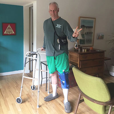 Scott flashes a peace sign with his fingers, while standing in his home and holding a walker with his other hand.
