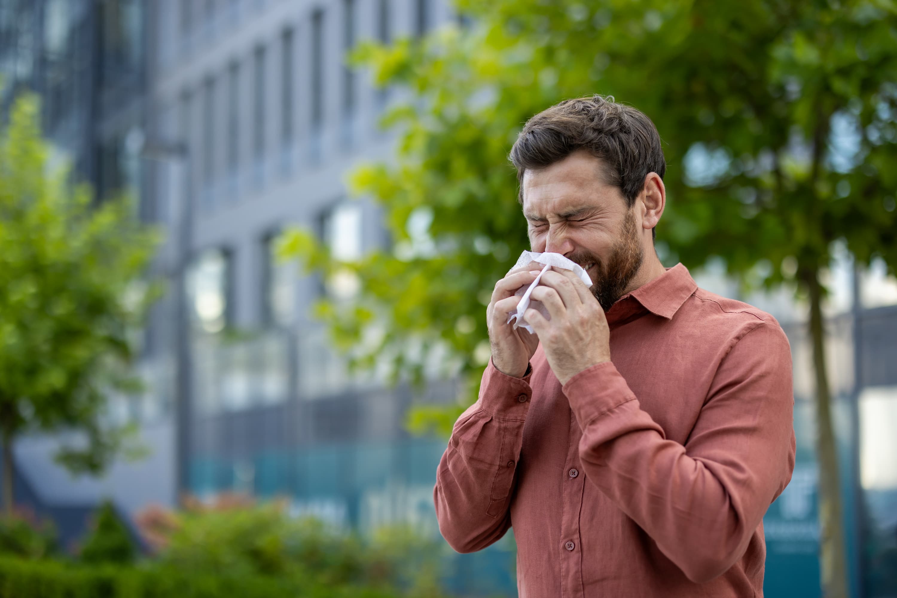 A man holds a handkerchief to his nose as he sneezes while standing outdoors.