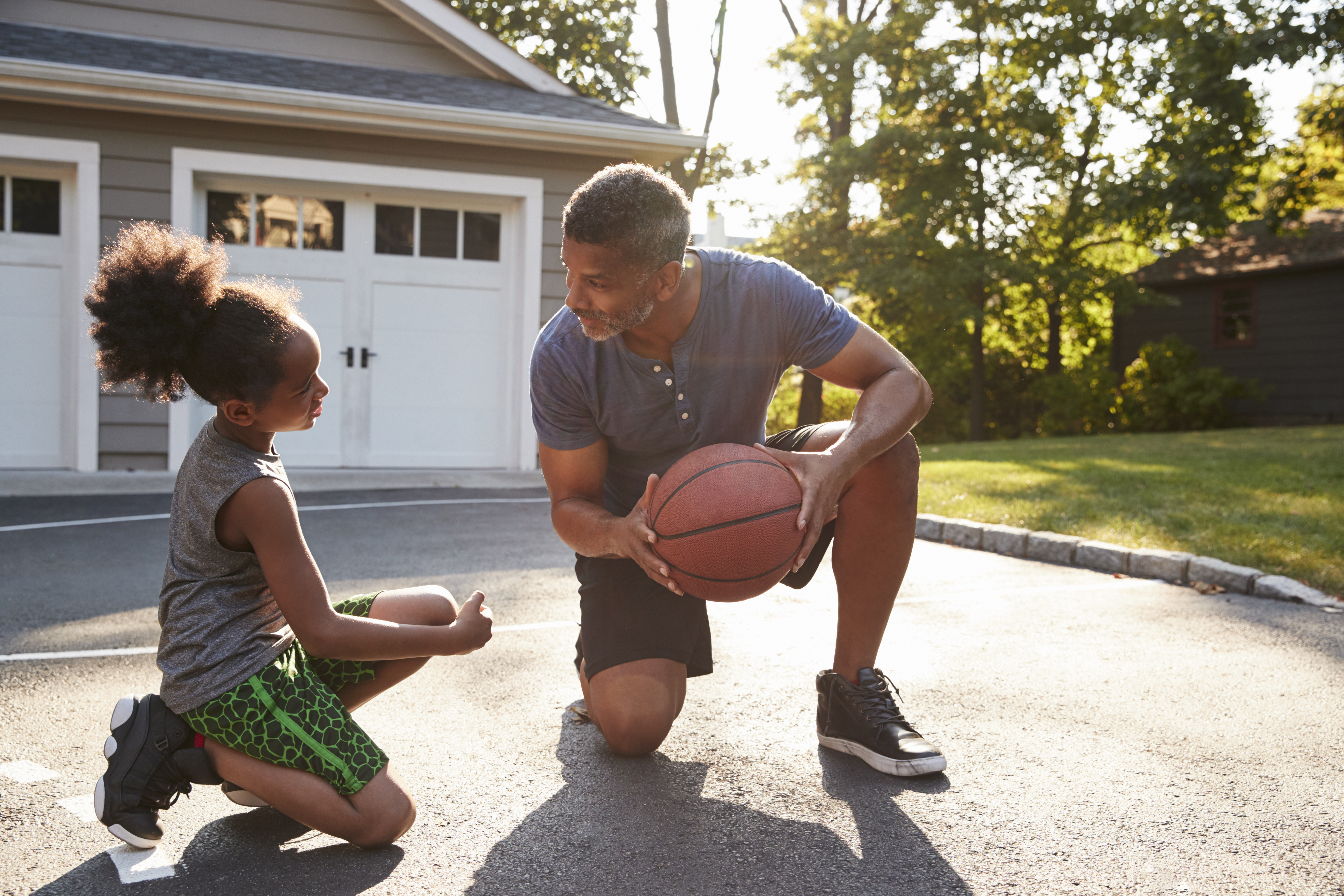 A man  holding a basketball speaks with young girl outdoors on a driveway