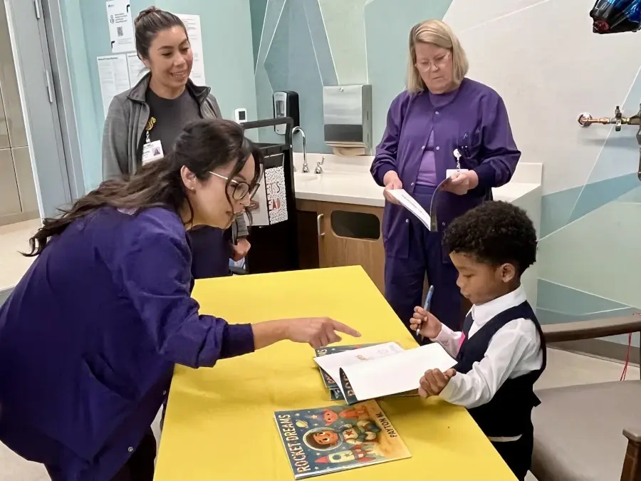 Young boy Payton seated at table with books surrounded by female hospital staff.
