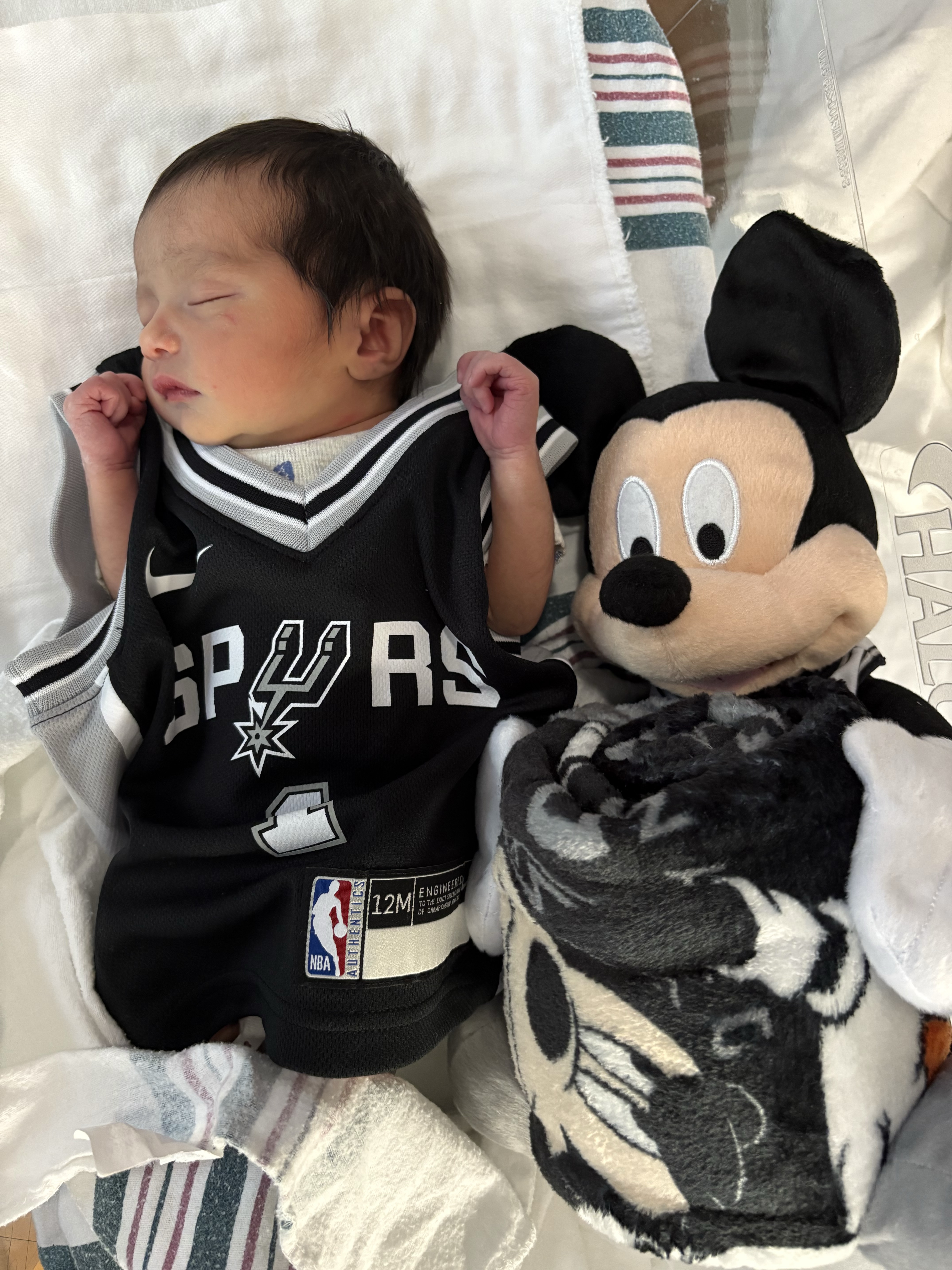 Newborn baby in hospital crib, wearing a spurs jersey and a spurs plush toy.