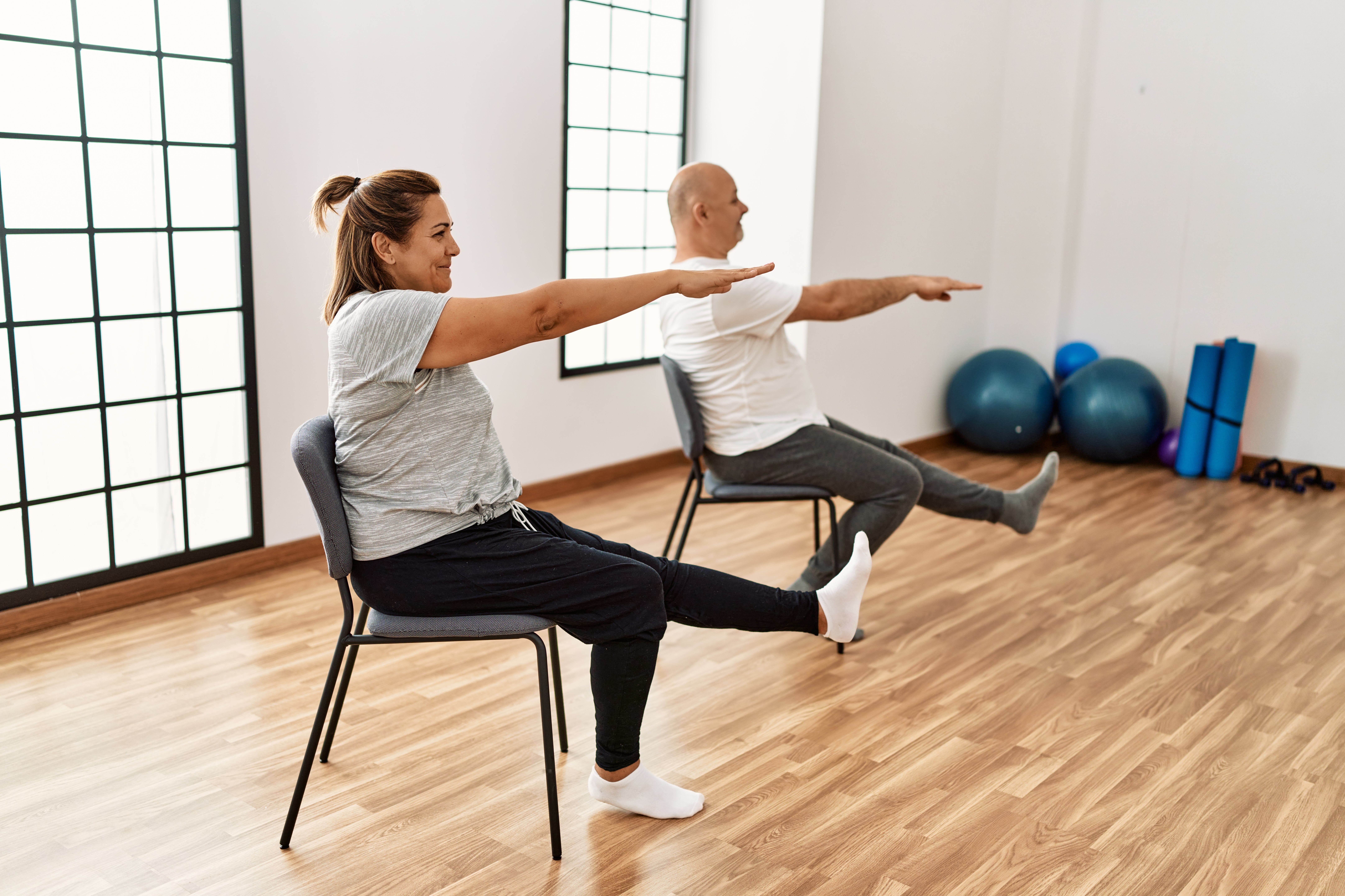 Middle aged woman and man holding yoga poses while seated on chairs in a workout room.
