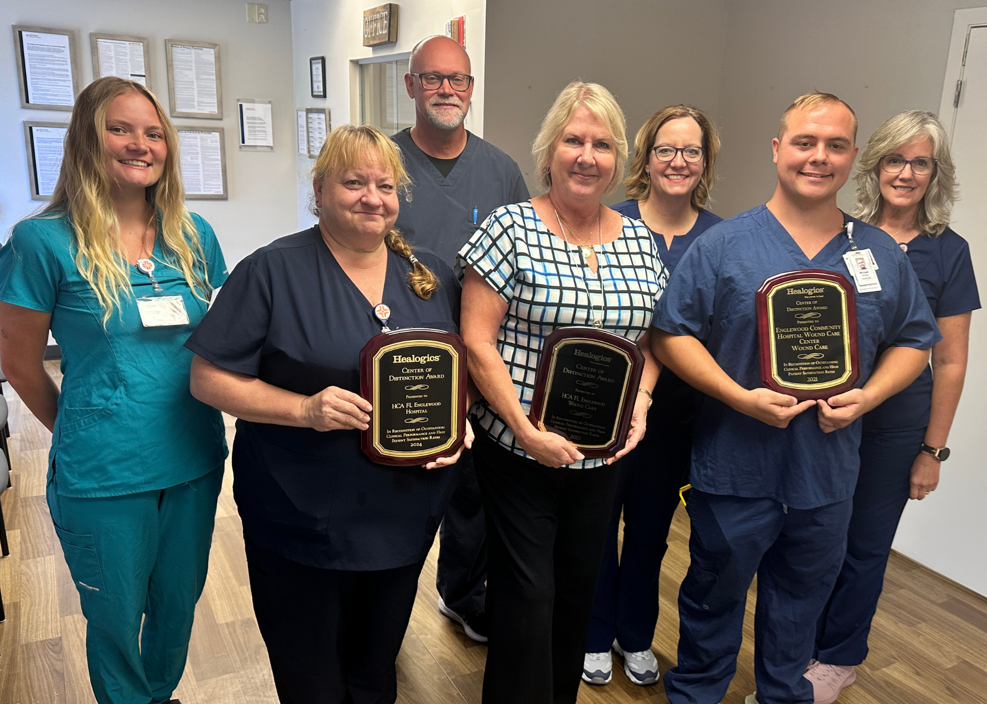 A group of seven people, all wearing medical scrubs or professional attire, stand together in a room. Three individuals in the front row are holding plaques that read "Center of Distinction Award". 