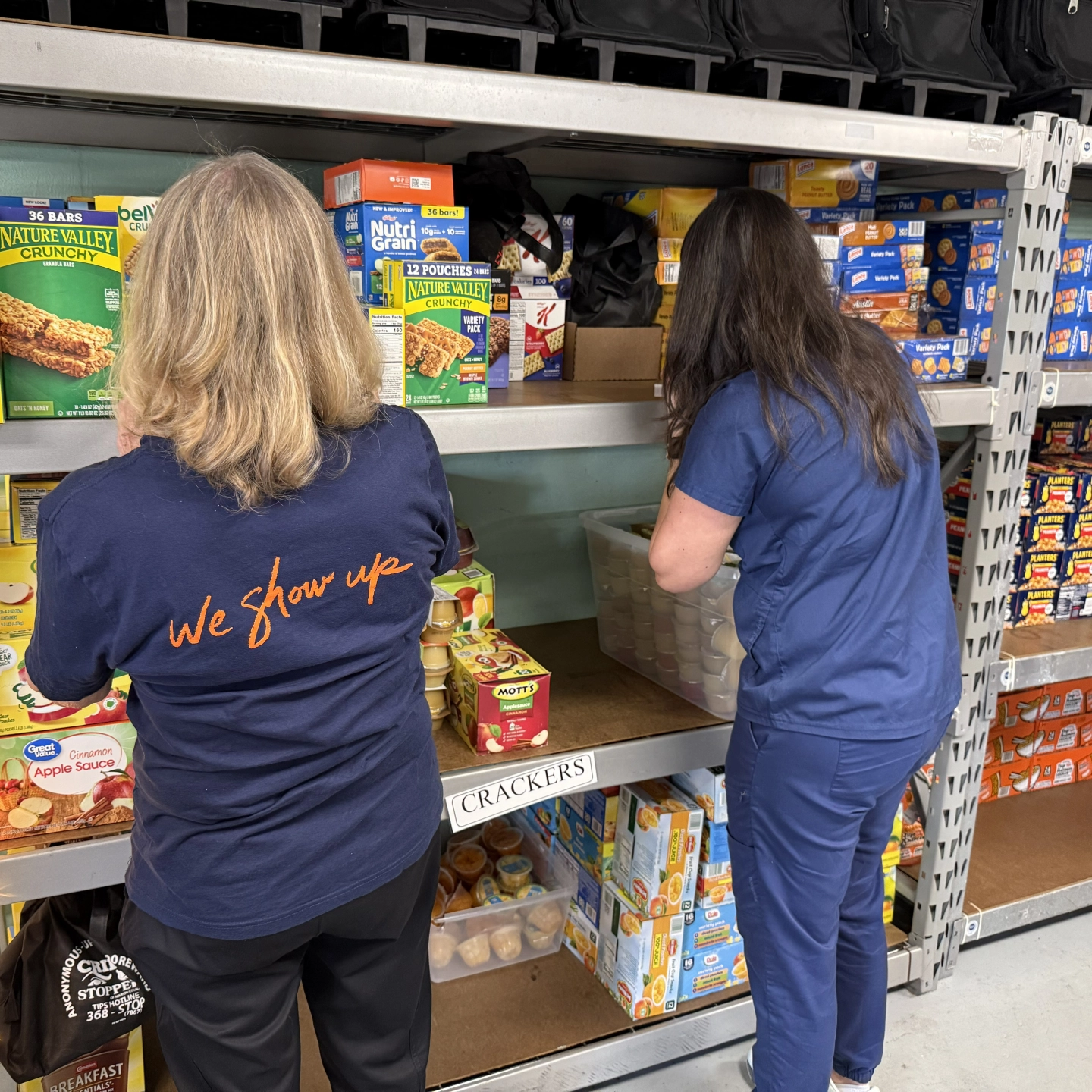 Two women in hospital scrubs organize a shelf of boxes of food.