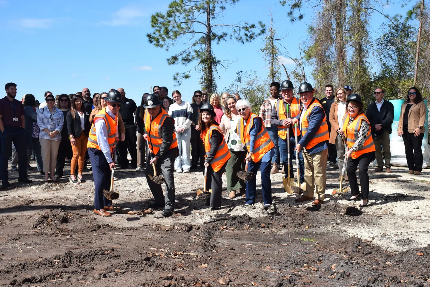 Several people wearing orange vests and hard hats holding shovels in the ground sounded by a group of people watching them.
