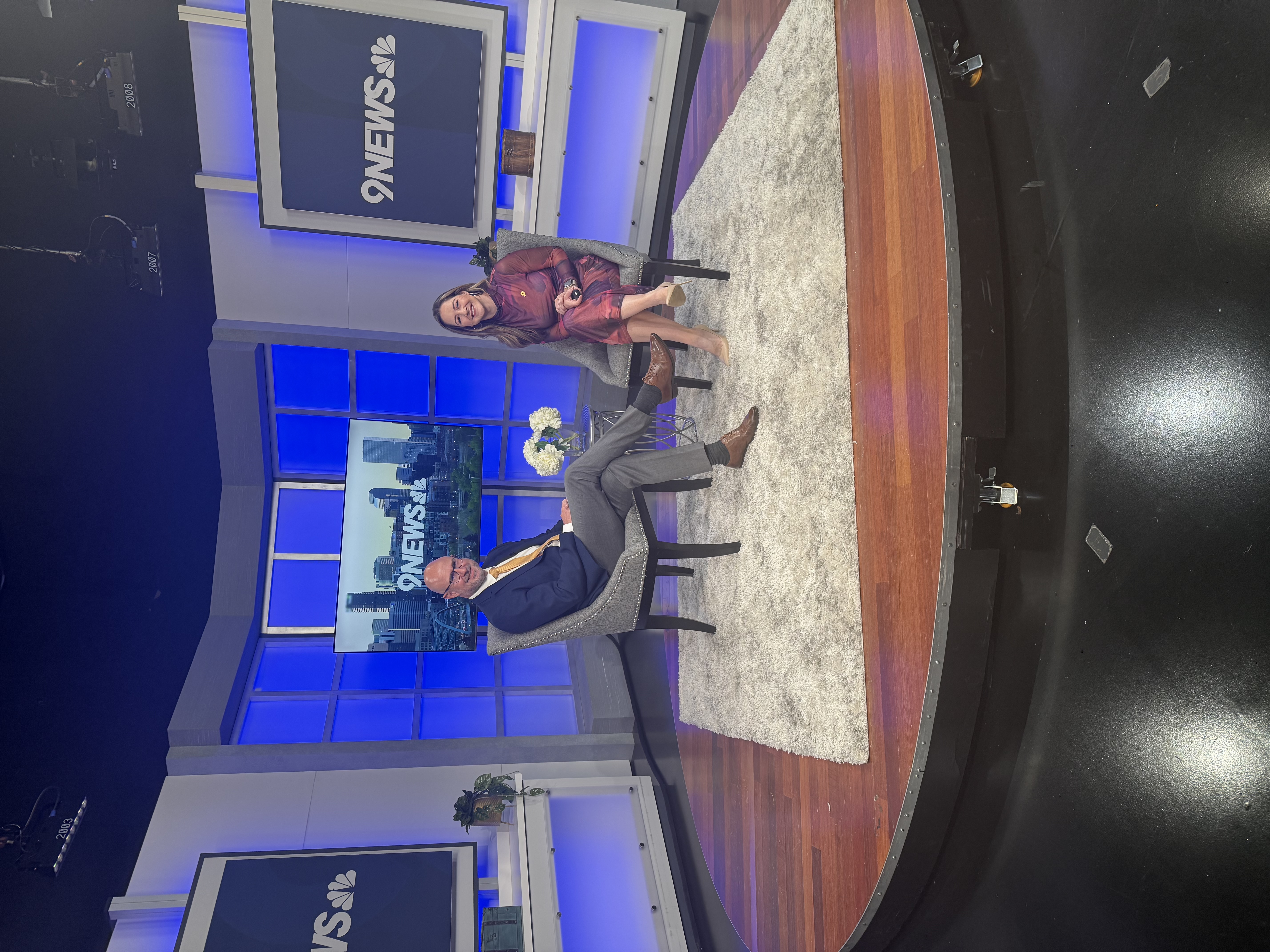 Dr. Warren Strutt and a news anchor are both seated on the 9NEWS studio set, smiling at the camera with the Denver skyline displayed behind them.