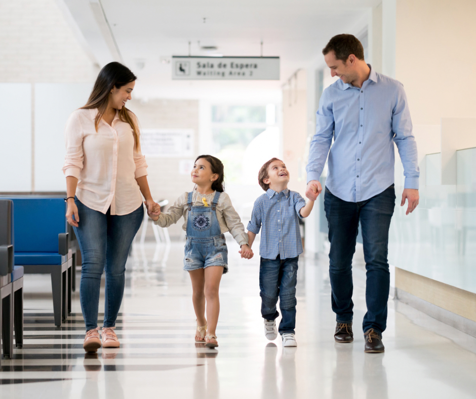 A family of four  - adult male and female, walking with two children - holding hands walking in a hospital hallway