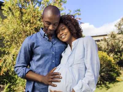 Standing outdoors, a man rests his hand on his smiling, pregnant wife's baby bump.