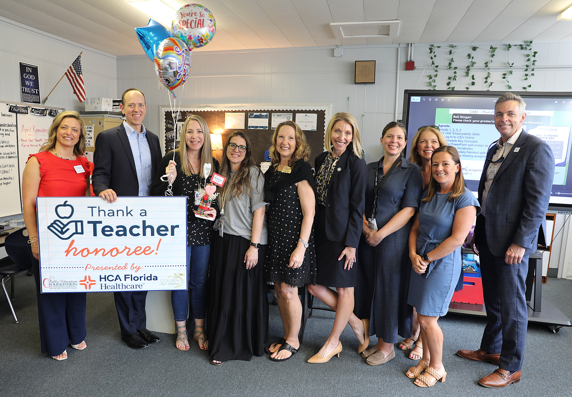A group of educators and staff stand together in a classroom posing for a photo. One person holds a "Thank a Teacher honoree!" sign while another holds balloons. A smartboard, posters, and classroom decorations are visible in the background.