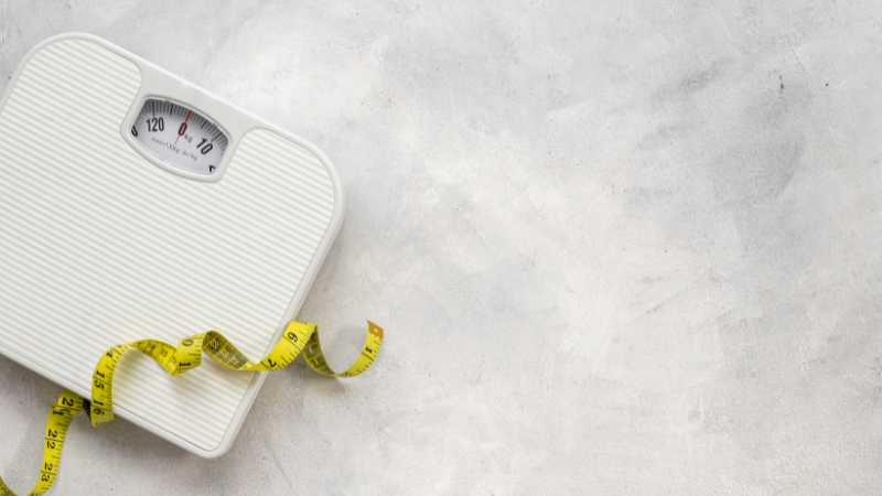 A white, body mass bathroom scale on the floor, with a yellow, coiled measuring tape draped over it.