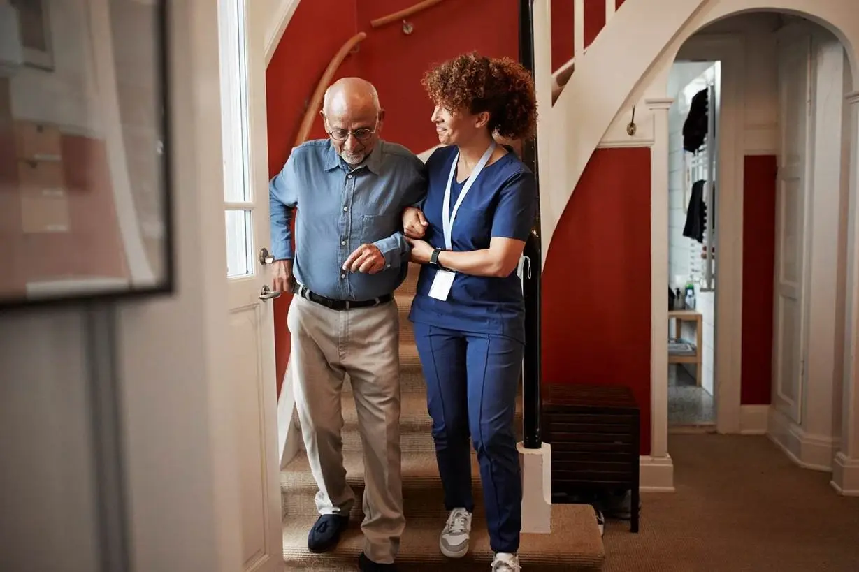 A nurse smiles while helping an elderly man walk down his home stairs.