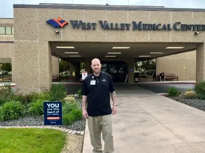 Surgery survivor David Riker wears a beard and hospital volunteer scrubs, standing outdoors.