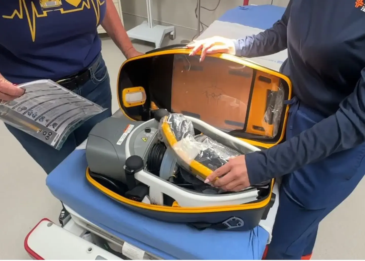 Two individuals stand beside a hospital bed examining an automated external defibrillator. One person holds an instruction manual while the other opens the device's case to reveal its internal components. The setting is an emergency triage room.