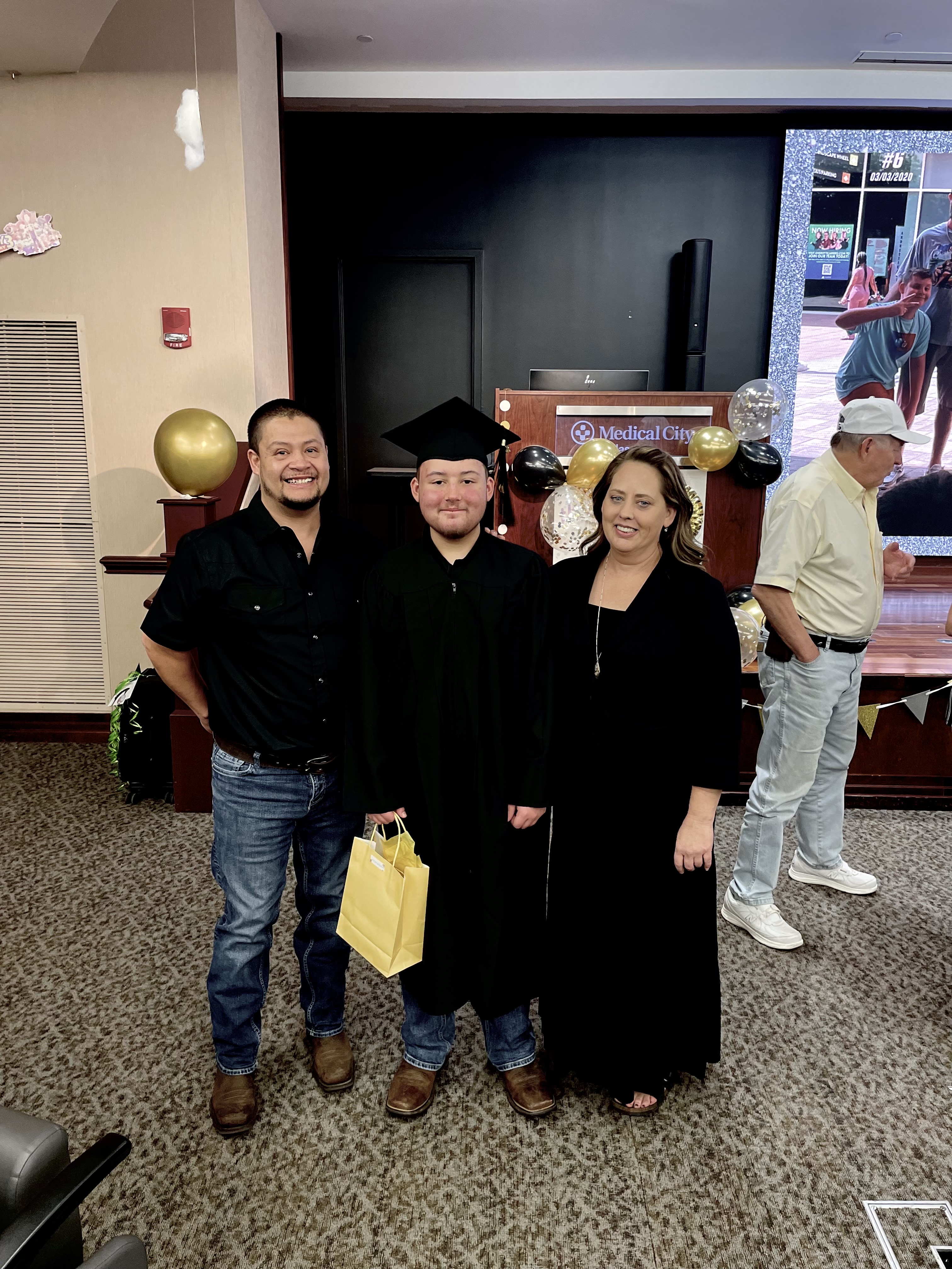 Tyler in a cap and gown standing between his parents as he celebrates his high school graduation at Medical City Children's Hospital.