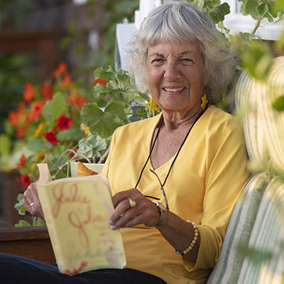 Mary Jane smiles for the camera, while seated on a patio chair outdoors, with plants behind her and a book in her hands.