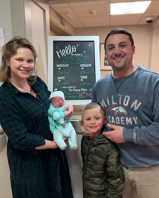 A smiling mom holding baby with dad smiling and big brother in front of a sign that says 'hello my name is John'.