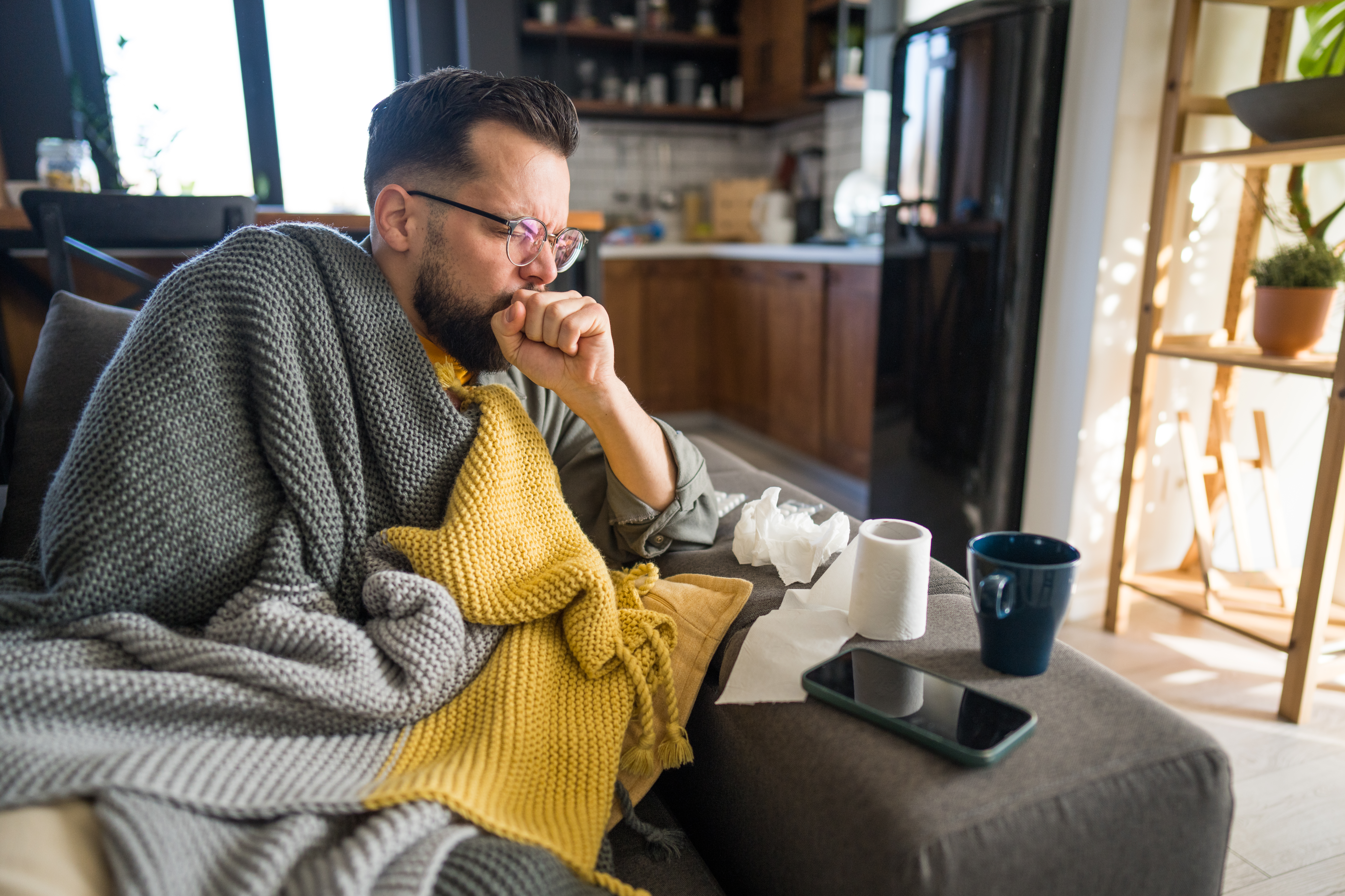 Sick man coughing on sofa at home, covered with blanket.