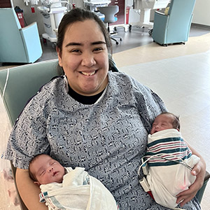New mother Chrystal B. smiles while holding her two newborns in either hand. 