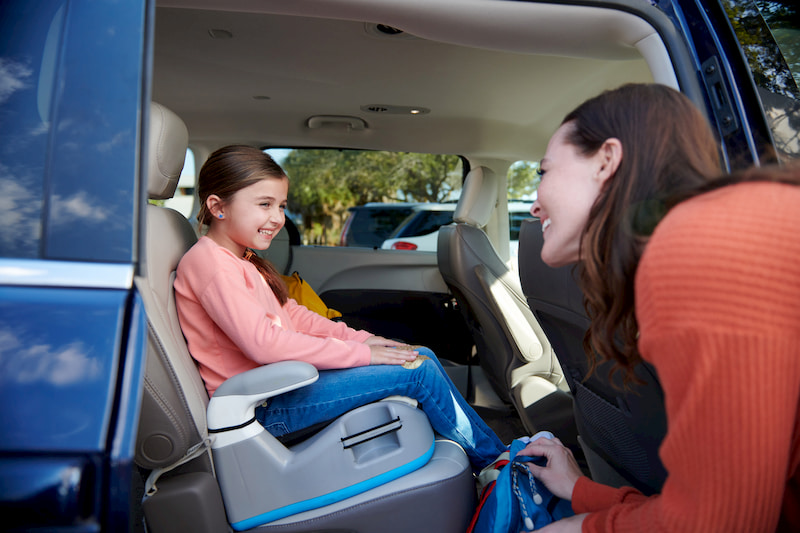 A young girl in a booster seat in a car smiles at her mother, who is placing a backpack in the car.