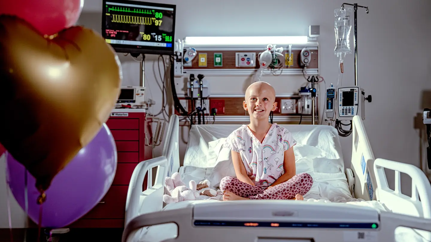 A pediatric cancer patient sitting on a hospital bed in a hospital room. Heart shaped balloons are in the forefront of the image and vital machines and equipment are in the background.