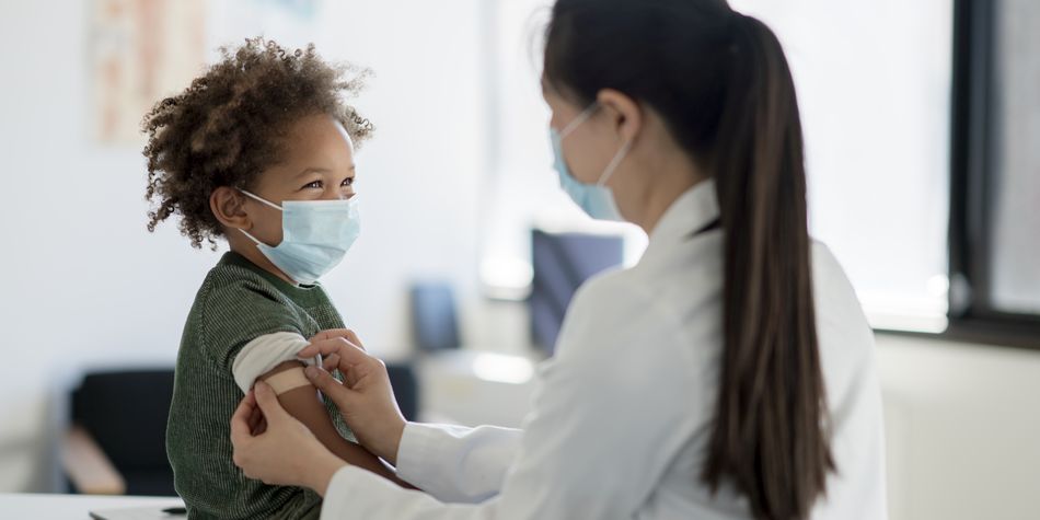 A young child sits up on an exam table as a doctor places a bandage on their arm after receiving a vaccine.