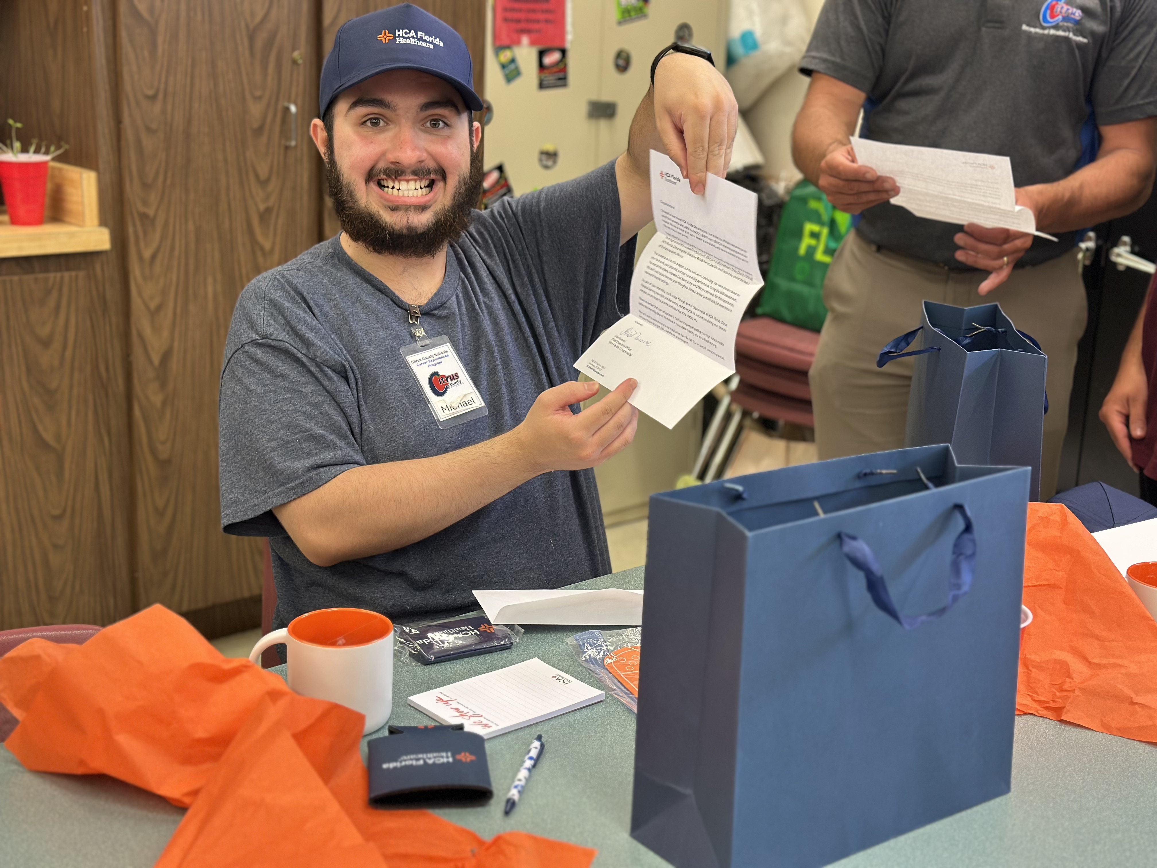 A student in an HCA Florida Healthcare hat smiles as he shows off an acceptance letter.