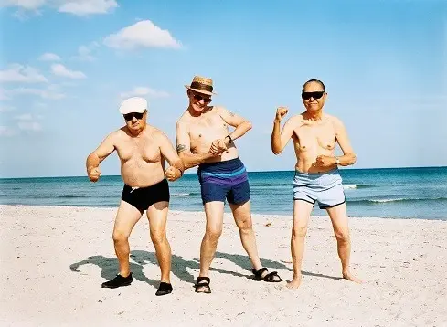 Three older men at the beach smile and flex their muscles.