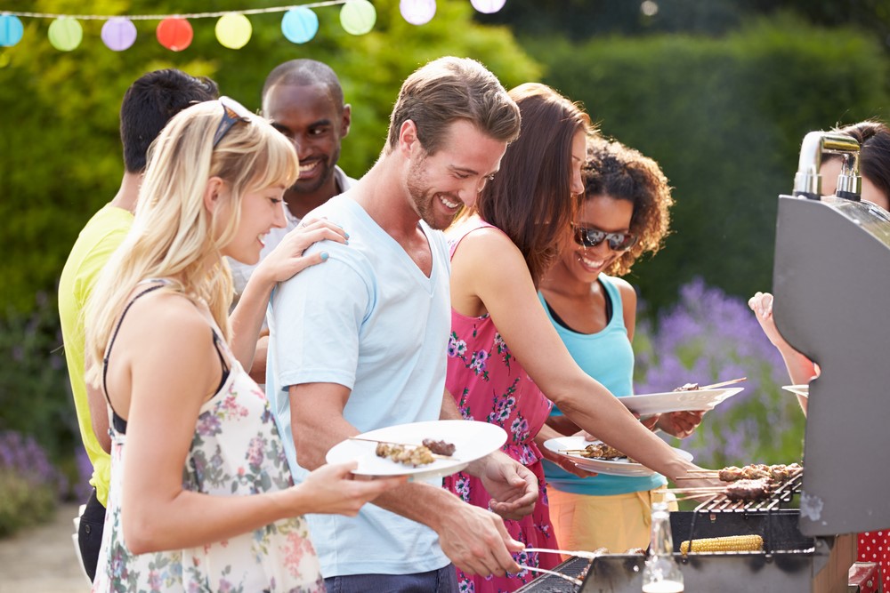 A group of smiling men and women at an outdoor party, filling their plates with food from a grill.