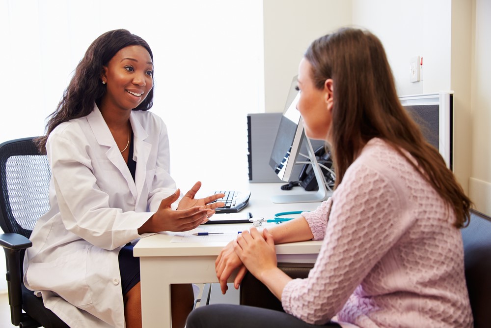 A physician speaks to her patient at a desk in an office.