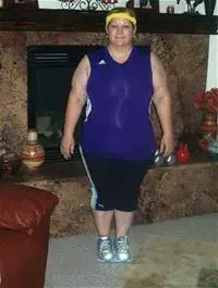 Debbie Searcy wearing a blue tank top, black workout pants and a yellow headband, standing in front of a fireplace.