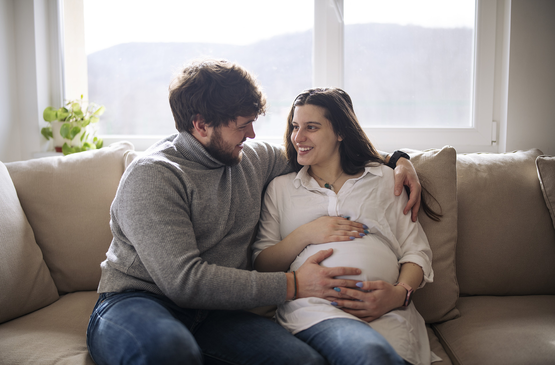 A couple sits together closely on a couch. The woman is expecting, and the man's hand is on her baby bump. They are both smiling.