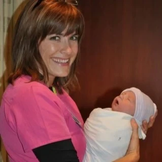 A female nurse in a pick-scrub top smiles while holding a newborn wrapped in a blanket and wearing a newborn hat.