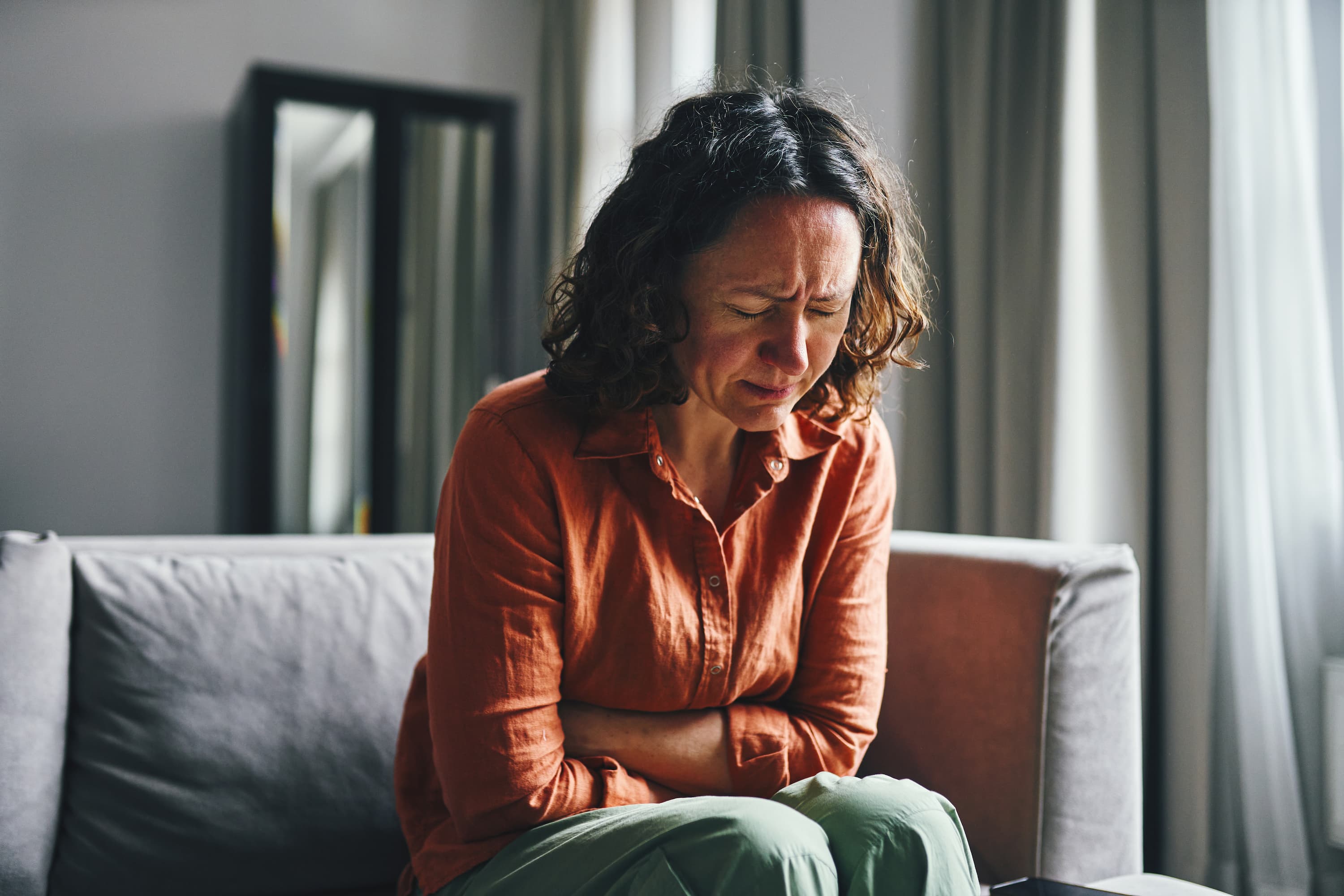 A woman sitting on a couch holding their abdomen and grimacing in pain, suggesting severe menstrual cramps.