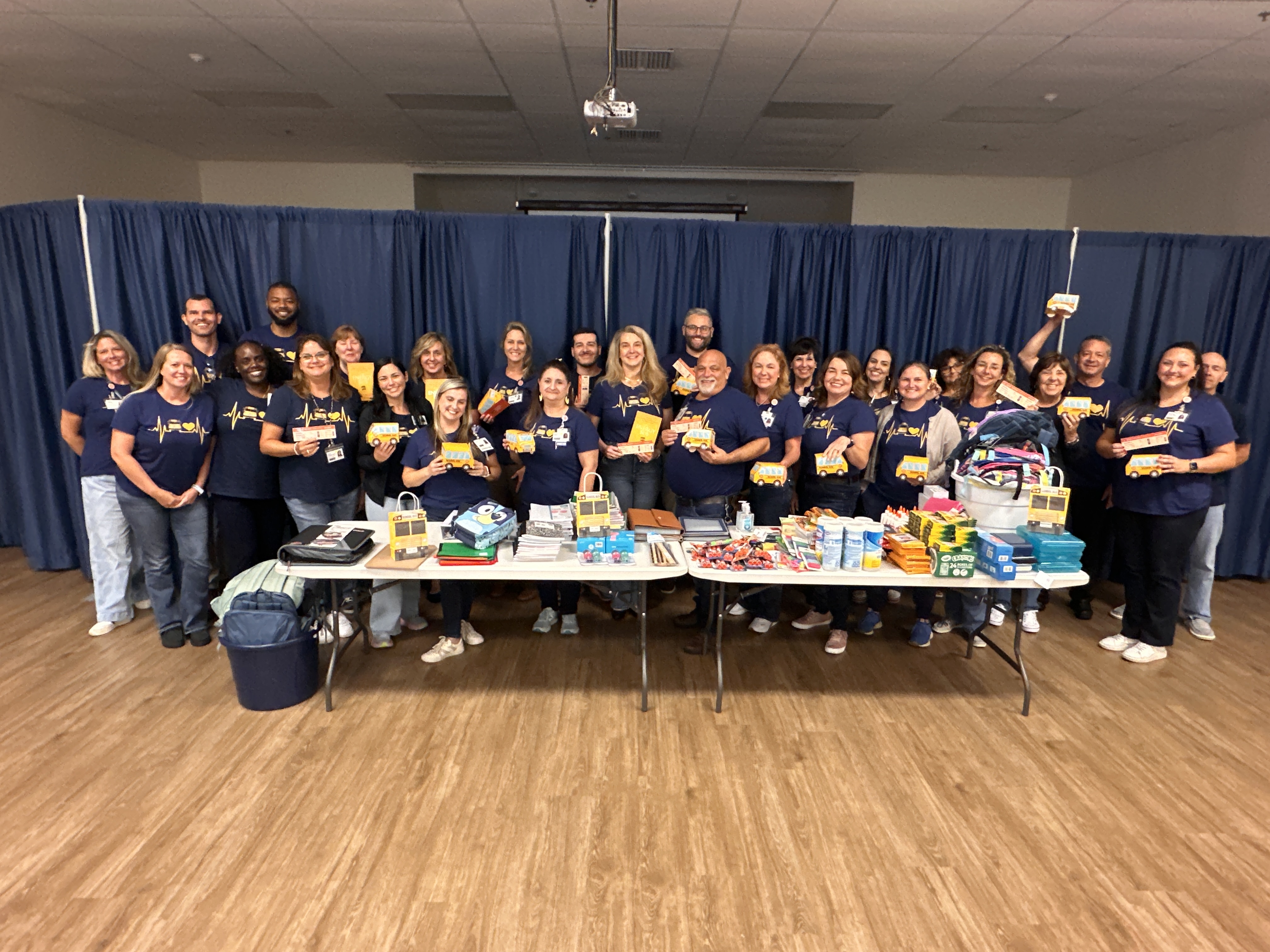 Around 30 people wearing matching navy blue shirts with a yellow school bus design stand behind two tables filled with supplies such as boxes, bottles, and bags. The group is posing for a photo in a large room. Some individuals are holding up items from the table.